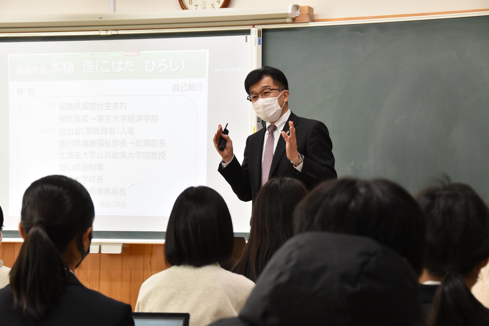 スクリーンの右側に立つ市長が身振り手振りで生徒たちに向かって話をしている様子の写真