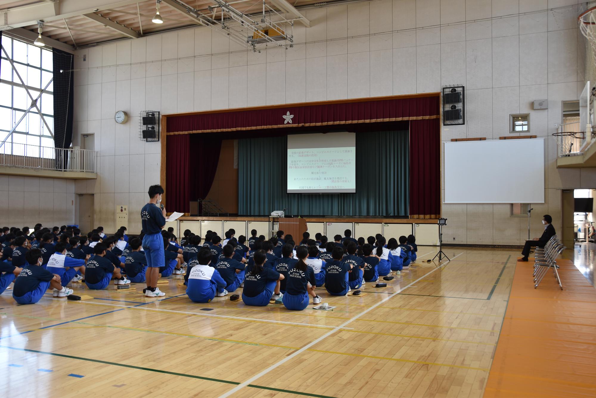 ステージに資料が映し出されたスクリーンが設置された、体育座りした生徒の中の男子生徒が立ちあがり質問を行っている様子の写真