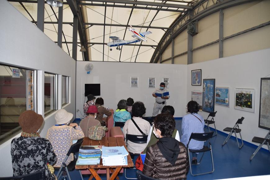額縁に入った資料が複数展示された白い壁で仕切られたスペースで、椅子に座る参加者に向けて、手に資料を持った職員の男性が話をしている様子の写真