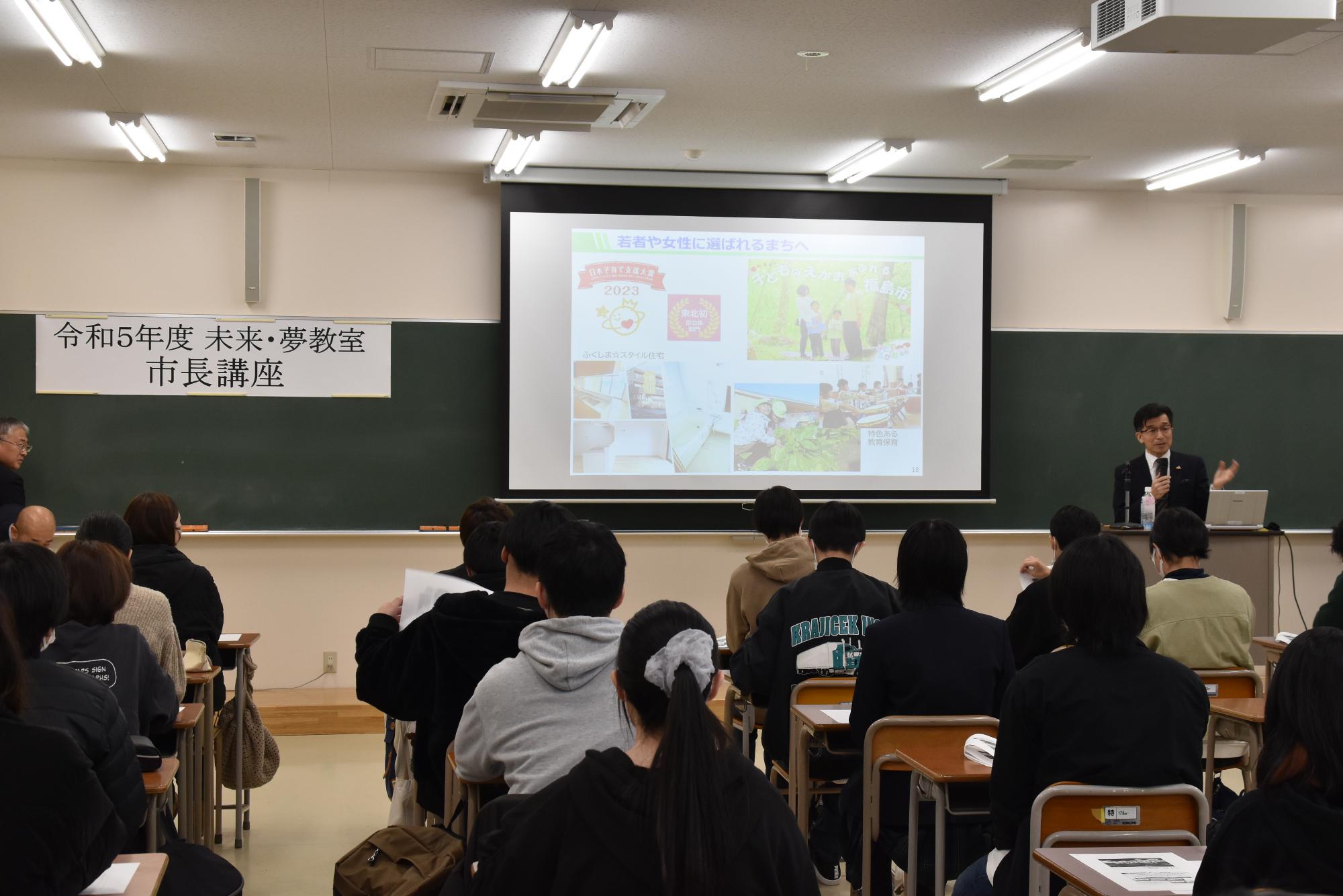 黒板の前に資料が映し出されたスクリーンが設置され、右側に立つ市長が話をしている様子の写真