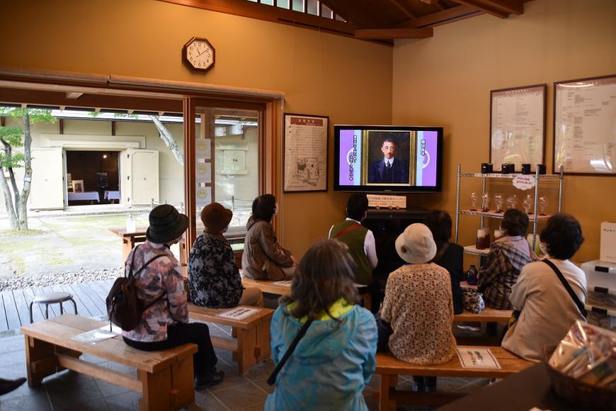 部屋の中に設置された木の長椅子に座った参加者たちが、モニターの映像を見ている様子の写真