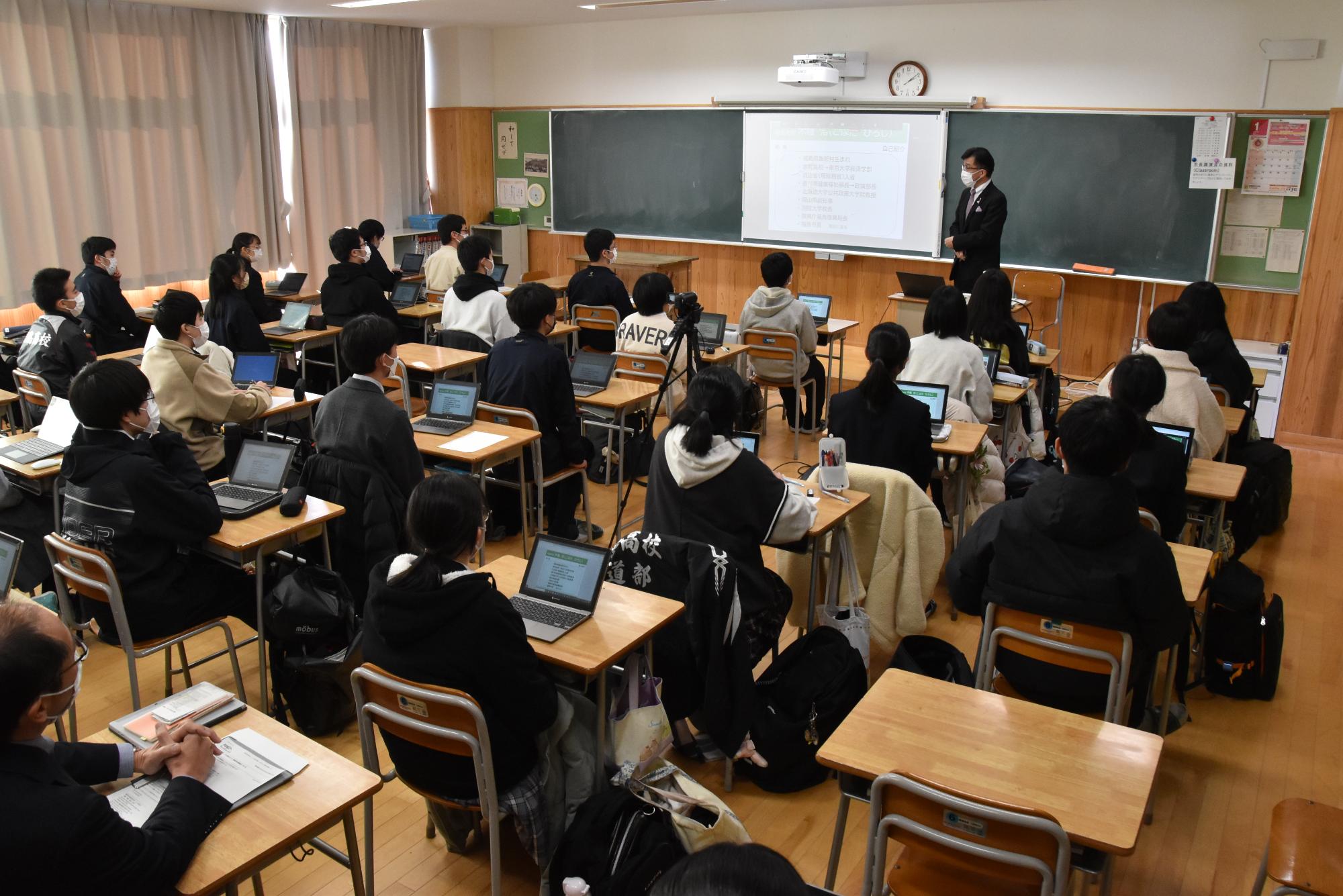 黒板中央に資料が映し出され、右側に立つ市長が生徒たちに向かって話を行っている様子の写真