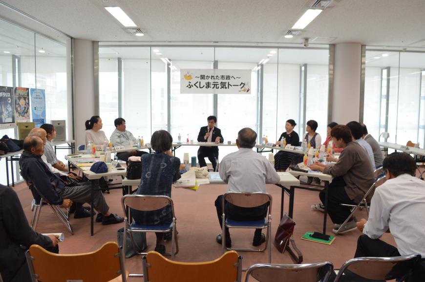 円形状に設置された机と椅子に座り、市長がマイクで話しをしており、参加者が注目して話を聞いている写真