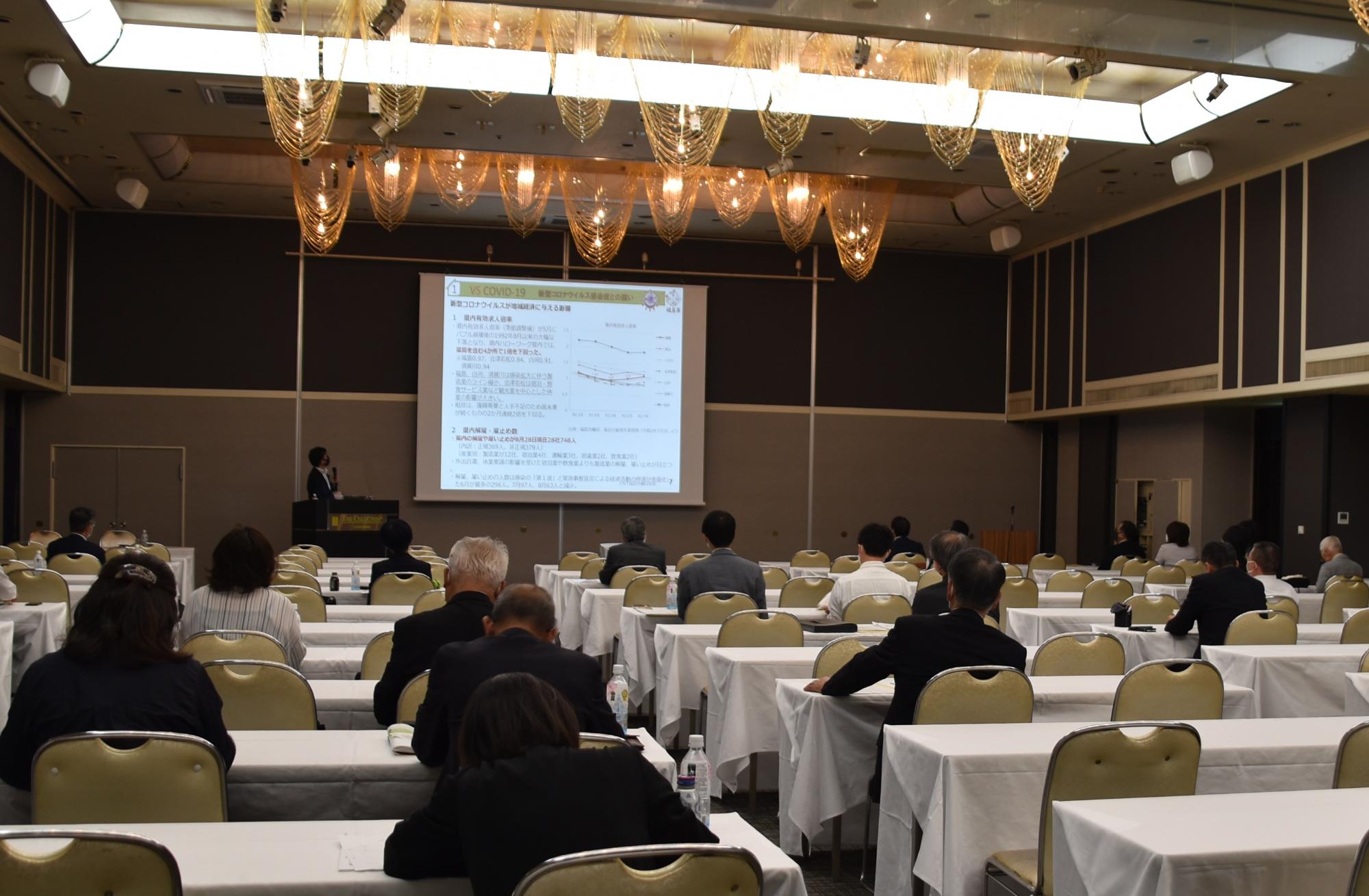 会場前方に資料が映し出されたスクリーンが設置され、左側の檀上に立つ市長が話をしている様子の写真