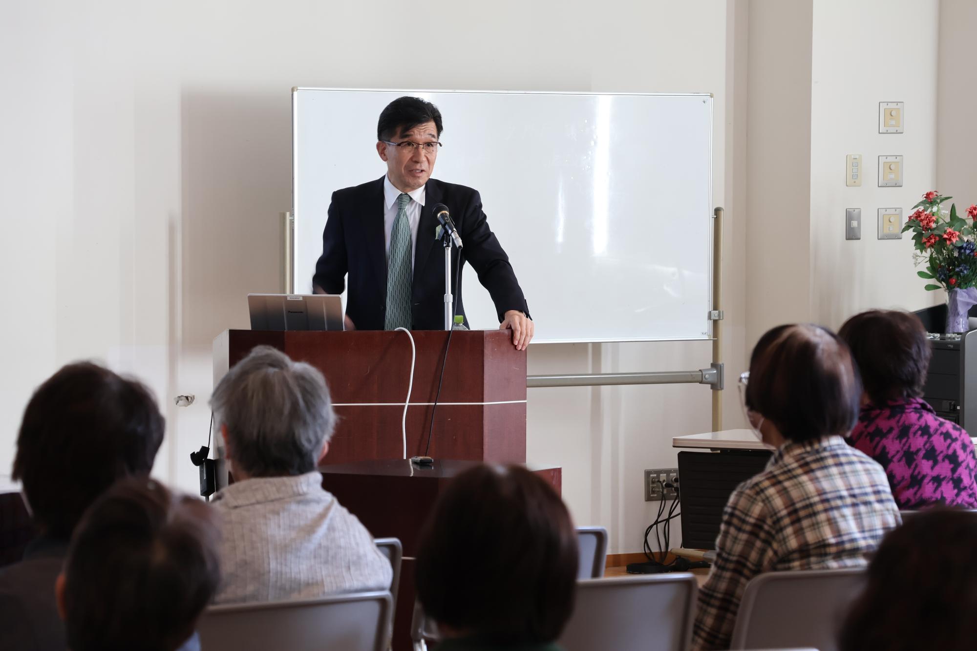椅子に座った参加者が聞いているなか、ホワイトボードの前に設置された壇上に立つ市長が話をしている様子の写真