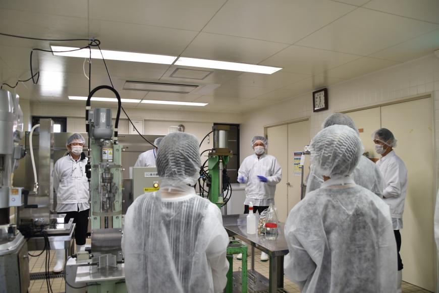 四季の里農産加工館内にある機械の前で係の方からの話を聞いている防護服を着た参加者の方々の写真