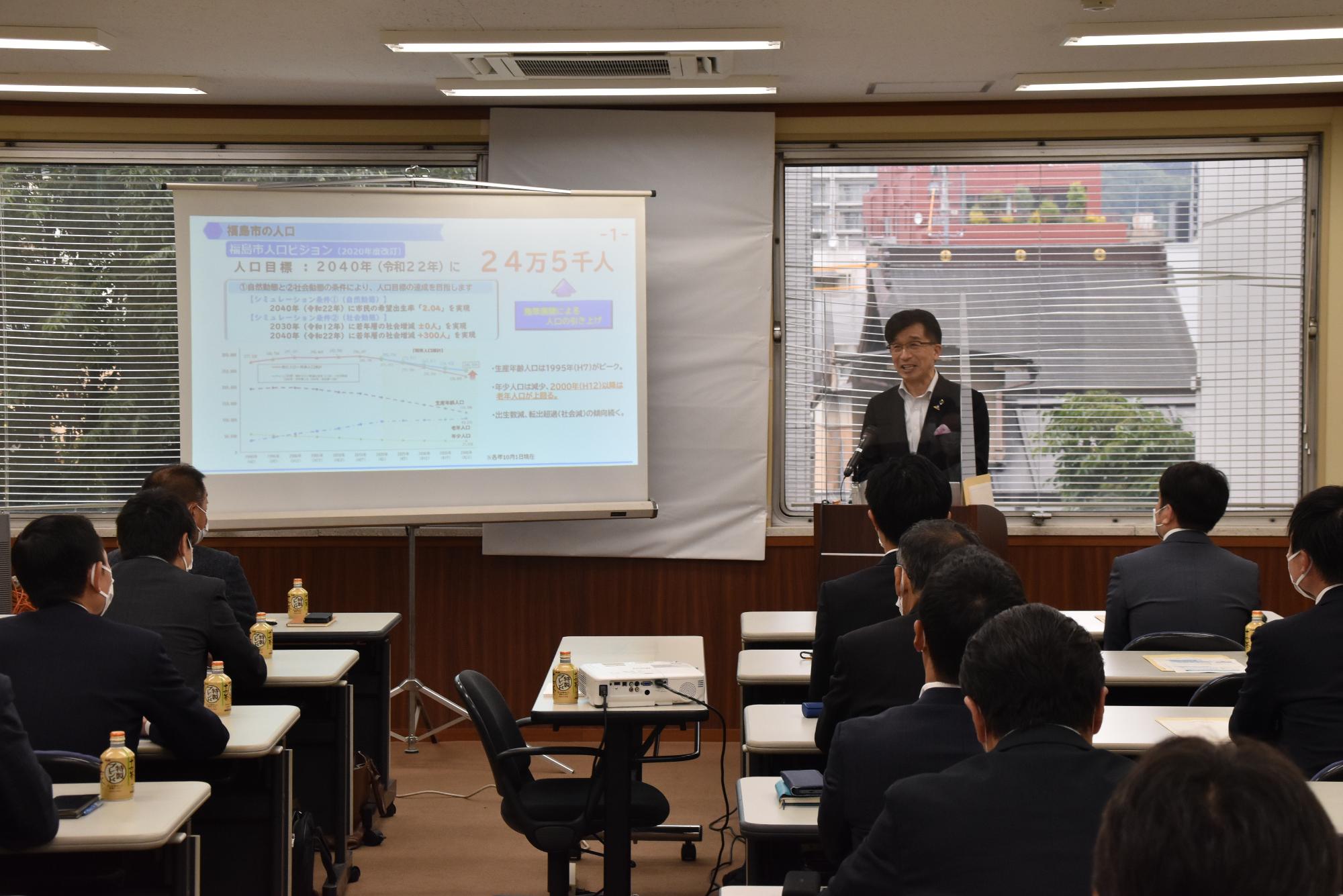 左側に資料が映し出されたスクリーンが設置され、右側の檀上に立つ市長が参加者に向かって話をしている様子の写真