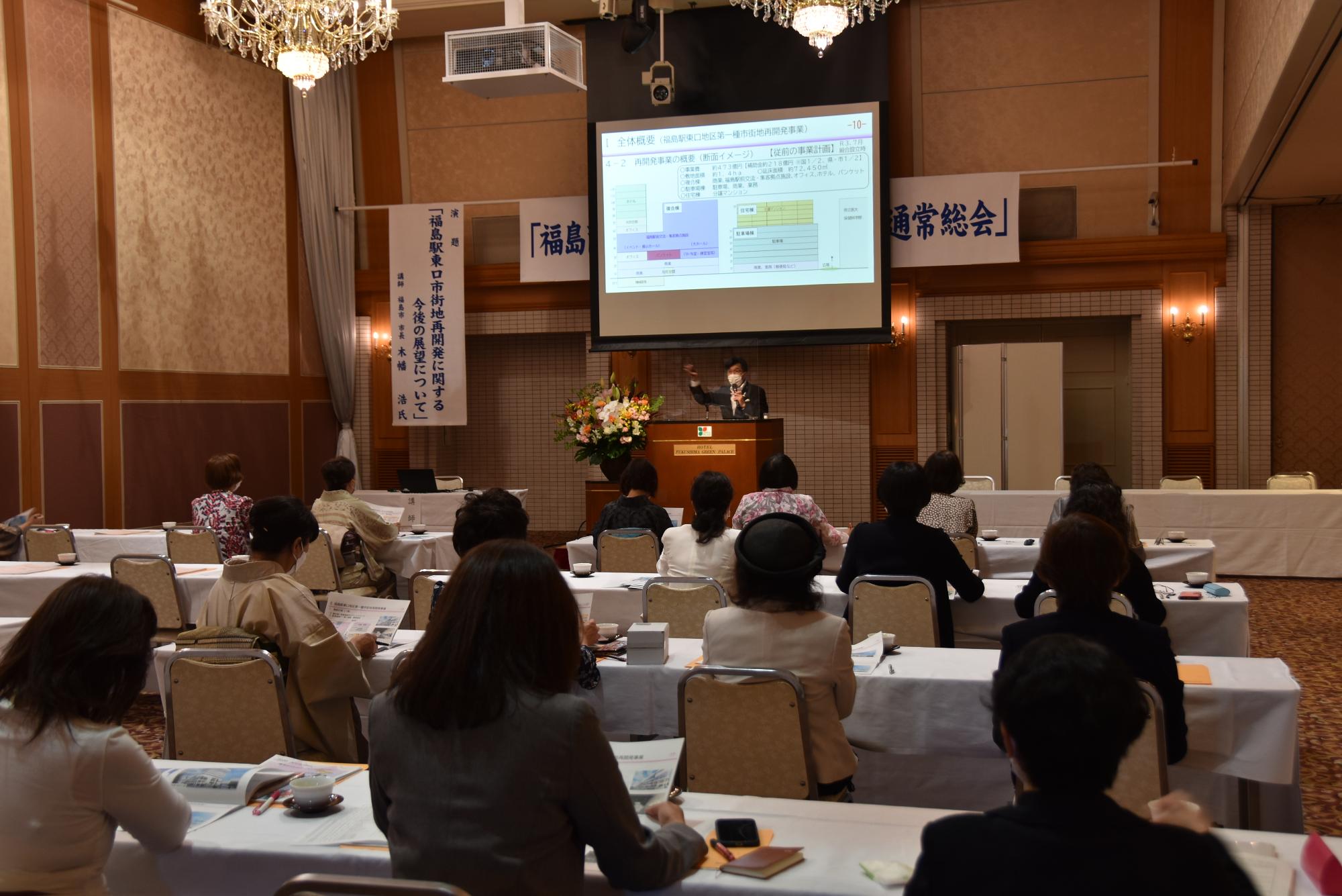 前方中央に資料が映し出されたスクリーンが設置され、檀上に立つ市長が参加者に向かって話をしている様子の写真