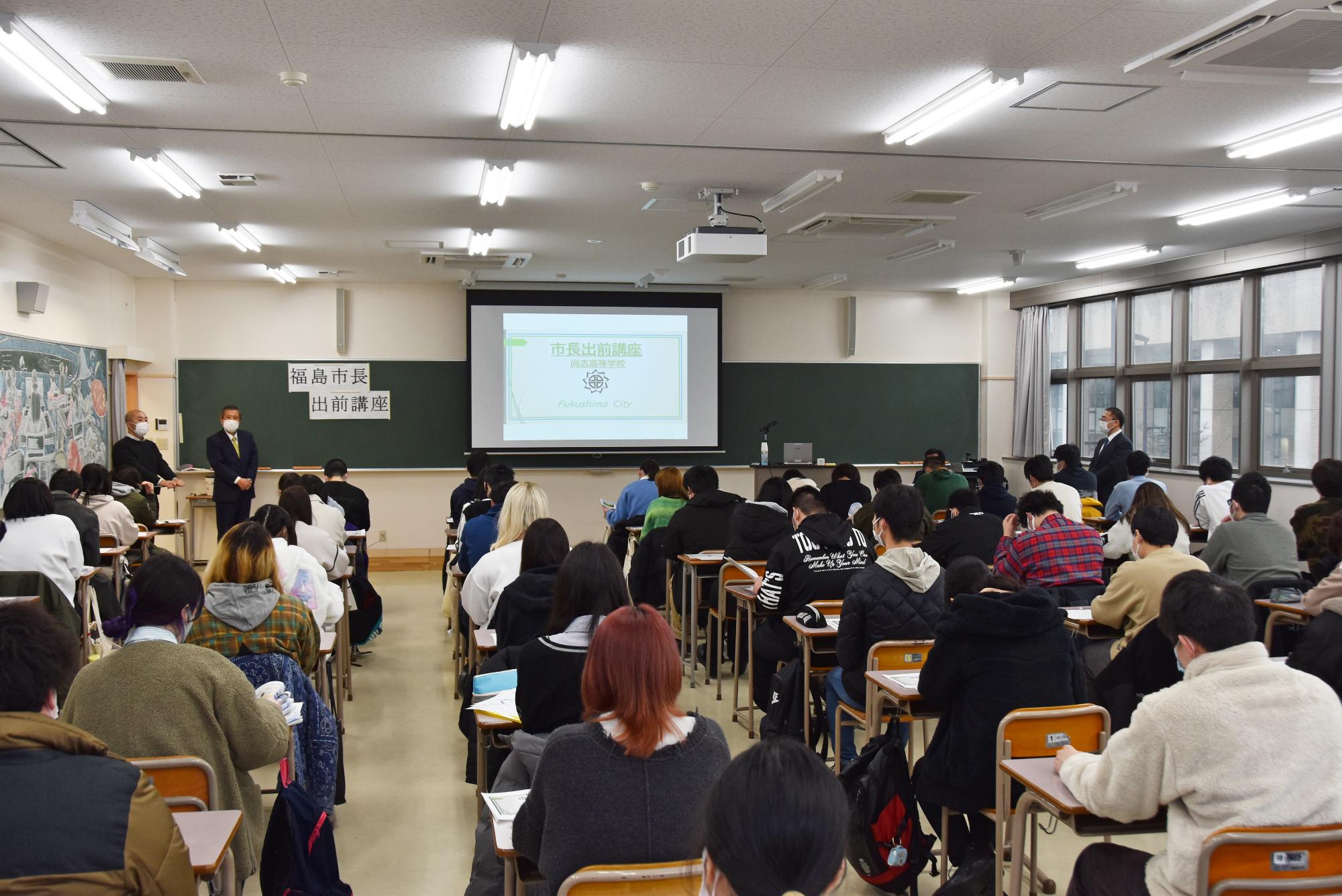 黒板の前に資料が映し出されたスクリーンが設置され、左側に2名の男性、右側に1名の男性が立つ教室内で講座が開始された様子を後方から撮影した写真
