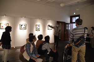 展示室に飾られた花の写真を関係者の方が説明している様子を椅子に座りながら参加者が聞いている写真