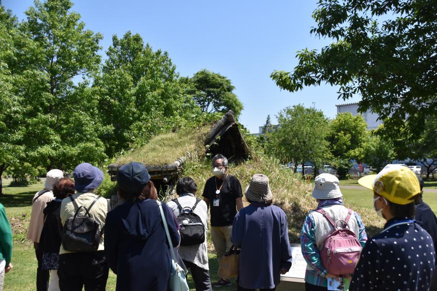 施設内に設置された建物らしき前で係の方が説明している様子を参加者が聞いている写真