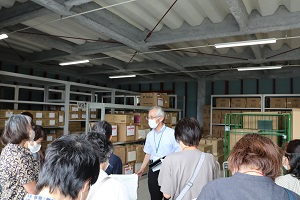 段ボールが棚に並んだ防災倉庫の中で関係者の説明を参加者が聞いている写真