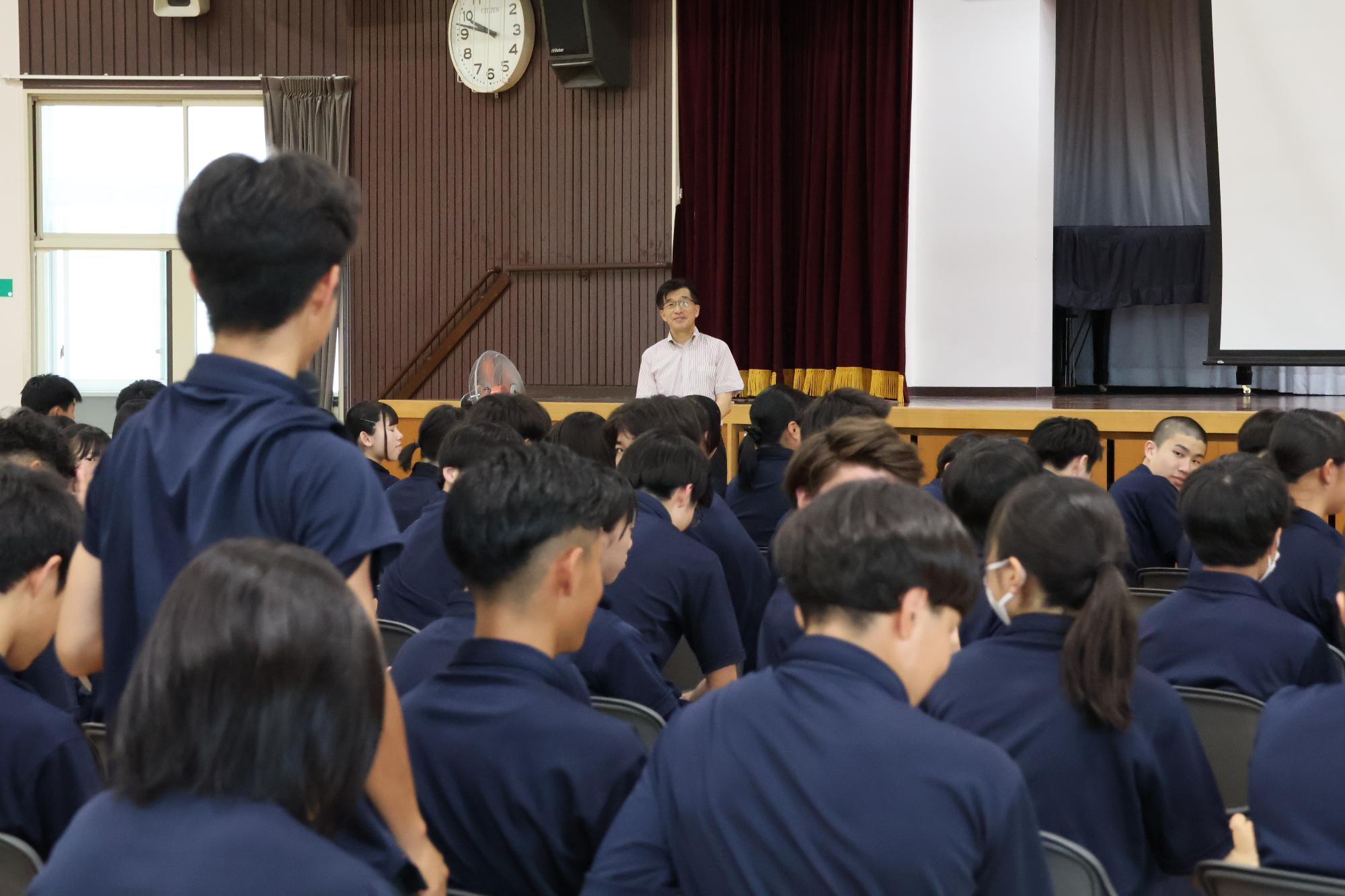 椅子から立ち上がった紺色のシャツを着た男子生徒が、市長に質問を行っている様子の写真