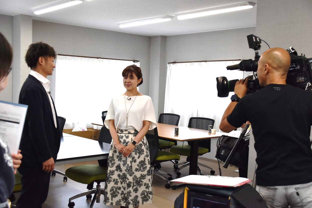 4脚ずつ椅子がセットされた2台のテーブルが並んだ室内で、カメラの前にスーツ姿の男性と女性アナウンサーが立っている写真