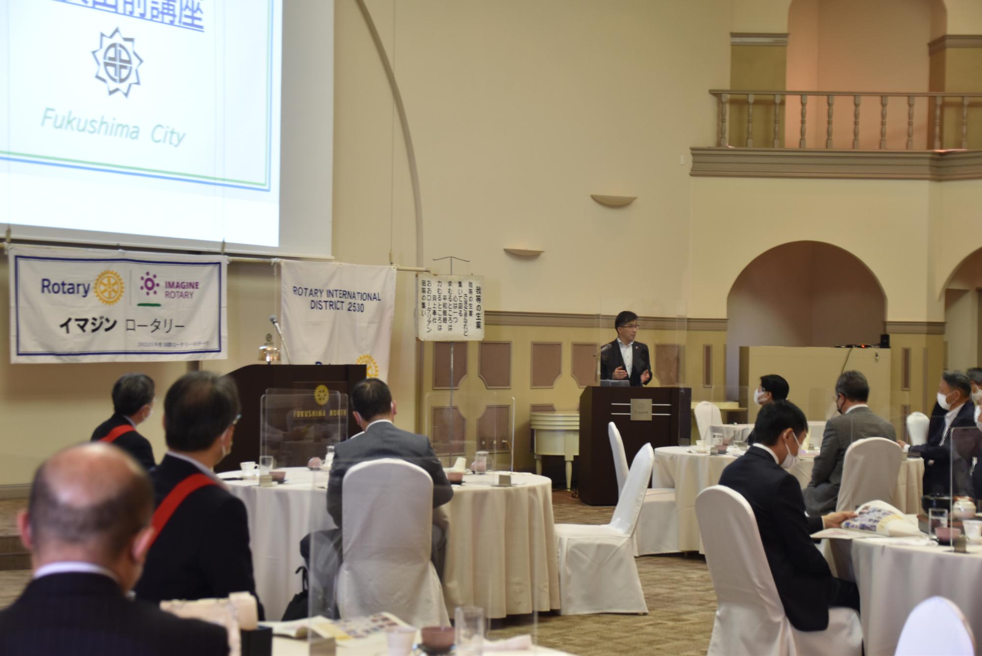 前方壁側に資料が映し出された大きなスクリーンが設置され、右側の檀上に立つ市長が話をしている様子の写真