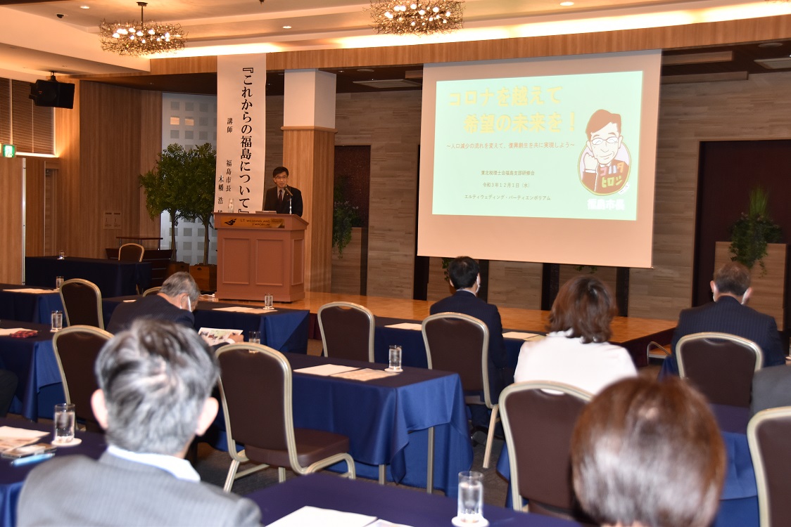 前方に資料が映し出されたスクリーンが設置され、左側の檀上に立つ市長が話をしている様子の写真
