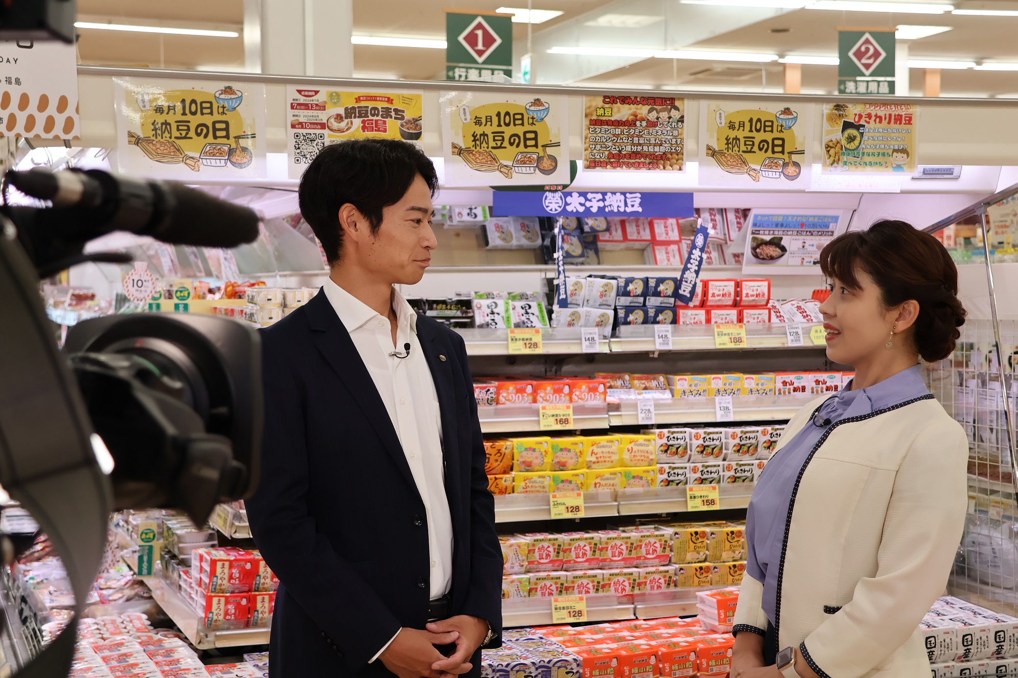 店内の納豆売り場の前で、スーツ姿の男性に女性アナウンサーがインタビューしている写真