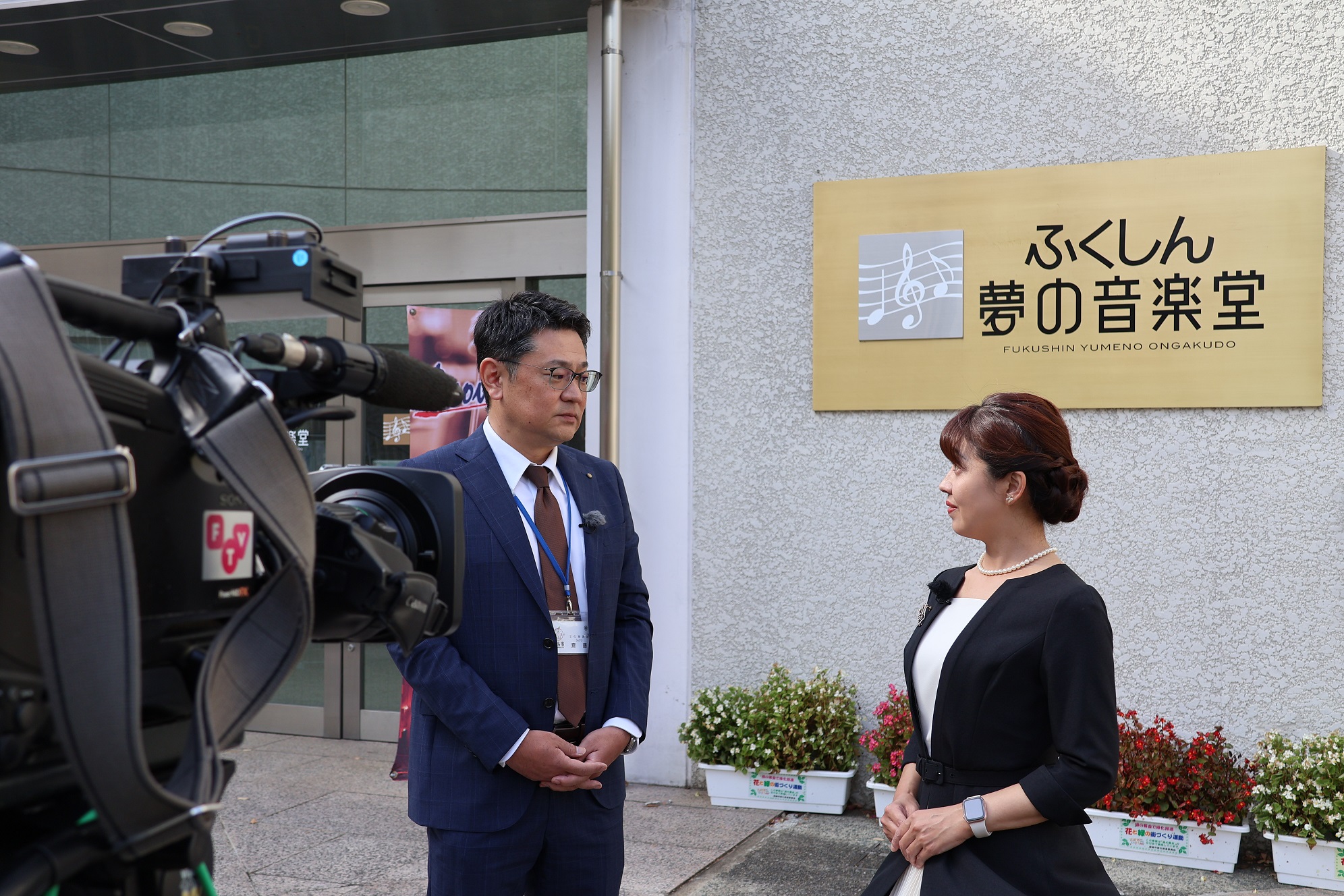 「ふくしん夢の音楽堂」の看板の前で、女性アナウンサーがスーツ姿の男性にインタビューしている様子の写真