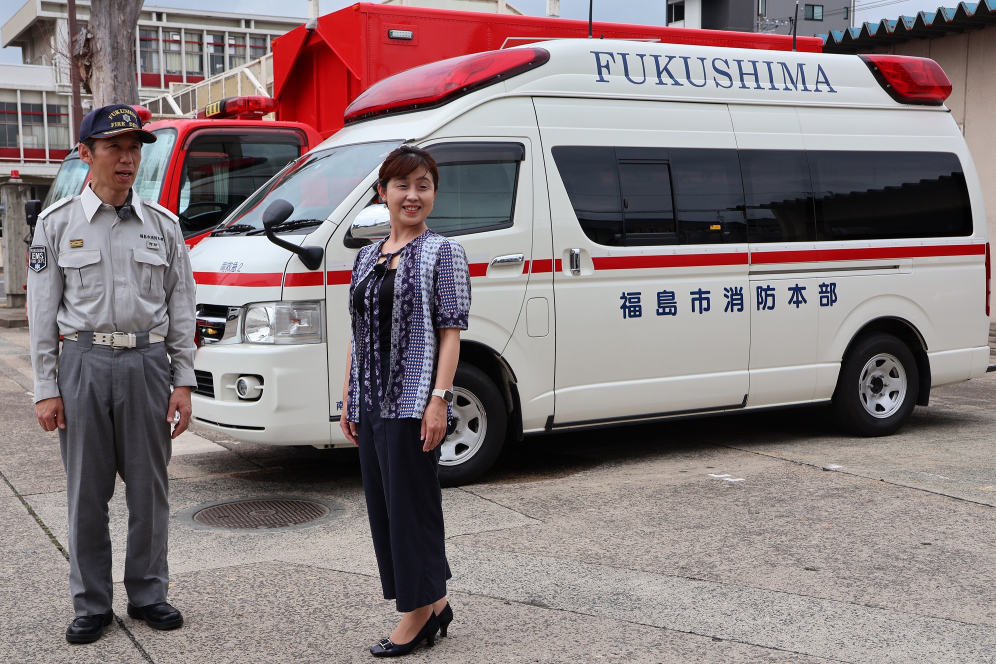 消防車の前で救急活動服を着た男性と女性アナウンサーが立っている写真