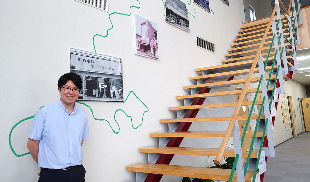 壁に「株式会社いちい」のモノクロ写真が展示された室内で撮影した秋山さんの写真