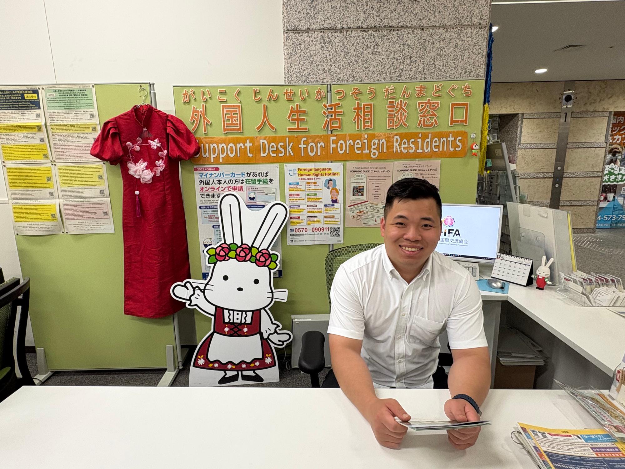 外国人生活相談窓口でクインさんが笑顔で座っている写真。ベトナムの民族衣装の赤いアオザイとももりんのパネルが飾られている。