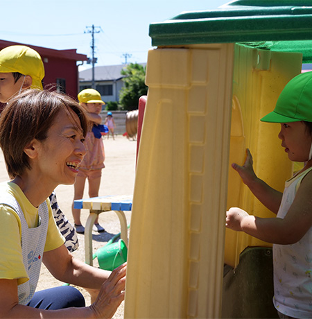 園庭に設置された小さな家に入った園児と遊んでいる笑顔の美智枝さんの写真