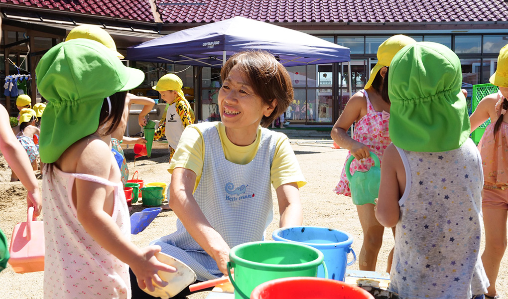 園庭でカラフルな色のバケツの前にエプロンを付けた美智枝さんが座り笑顔で園児と話をしている様子の写真