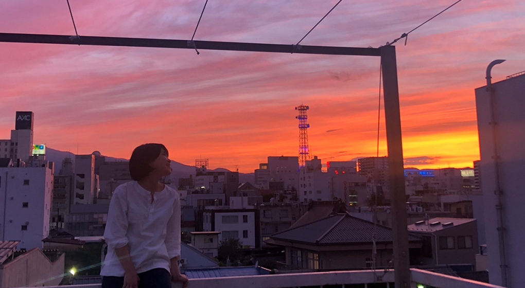 ベランダで赤く染まった夕暮れの空を見ている近藤 仁美さんの写真