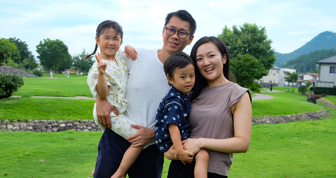 芝生が広がる公園内で旦那さんが女の子を抱っこし、美史さんが男の子を抱っこした紺野さん家族の写真