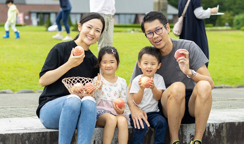 芝生広場の前に座る紺野さん家族が一人ずつ桃を持っている写真
