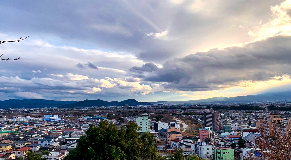 分厚い雲の隙間から太陽の光が差している空と福島市の街並みを高台から撮影した写真