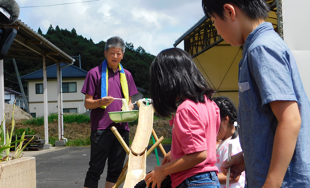 竹で作られた手作りのそうめん流しを子供達と楽しむ永井 康統さんの写真