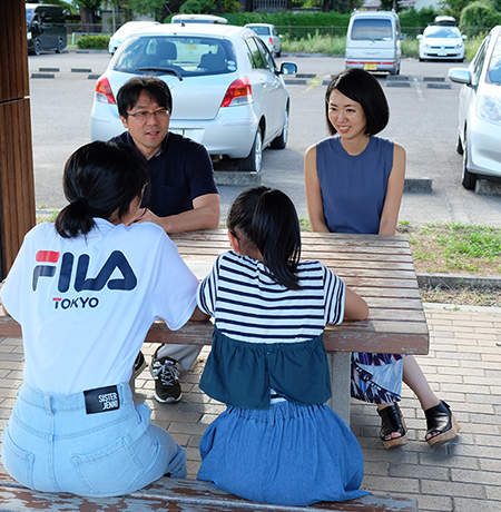 公園の木製テーブルが設置された休憩スペースで中島 明日香さん家族が座り話をしている様子の写真