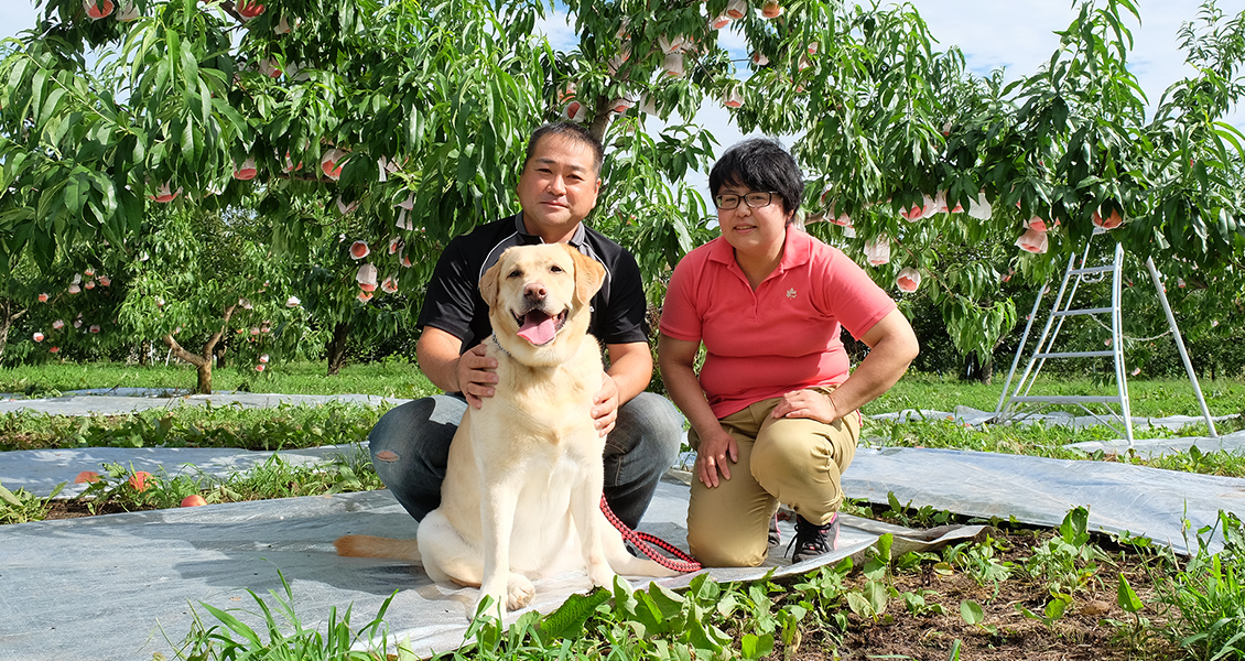果樹園を背景に大内さん夫婦と飼い犬の写真