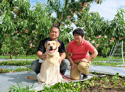 果樹園を背景に大内さん夫婦と犬の写真（大内 徹也/美千代さんのページへリンク）