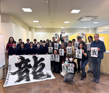 「龍」と書かれた大きな半紙を5人の女子生徒が持ち、右側に初めて書道を体験した外国人の方々がそれぞれ作品を持ち並んでいる写真