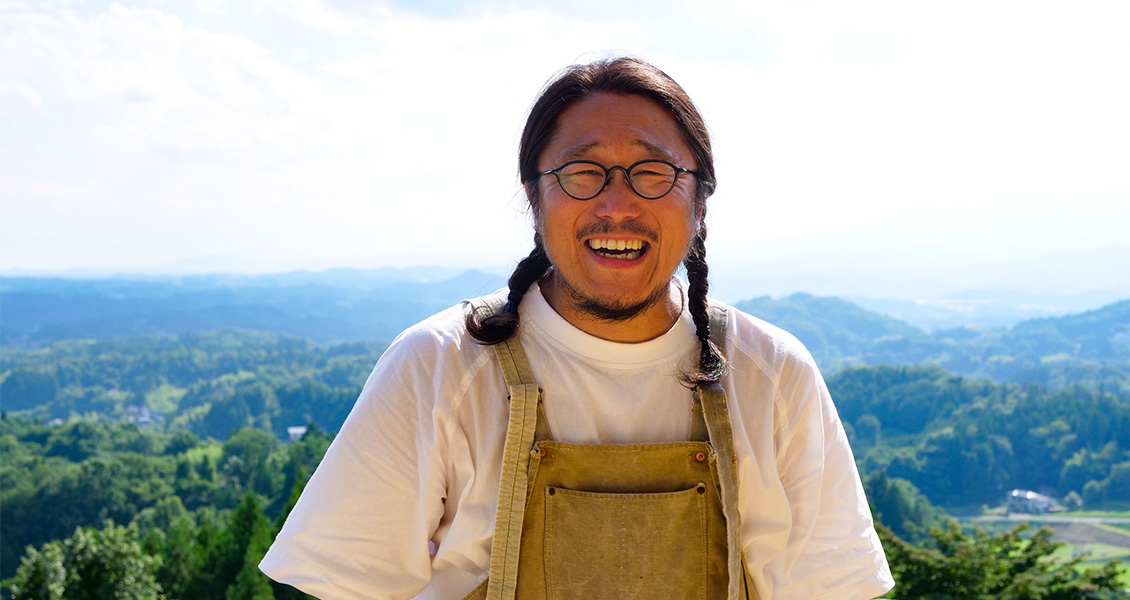 遠くの山並みまで見渡せる高台をバックに立つ笑顔の渡邊 浩志さんの写真