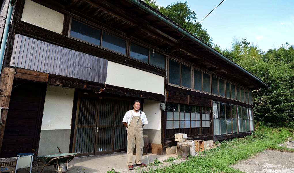 木々に囲まれた敷地に建つ、こげ茶色の外壁に沢山の窓ガラスが埋め込まれた古民家の前に立っている渡邊 浩志さんの写真