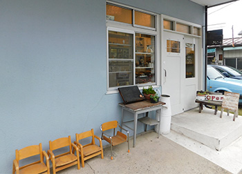 白いドアの左側に机と4脚の小さな椅子が並んで置かれた水色の外壁のきなこcafeの建物外観の写真