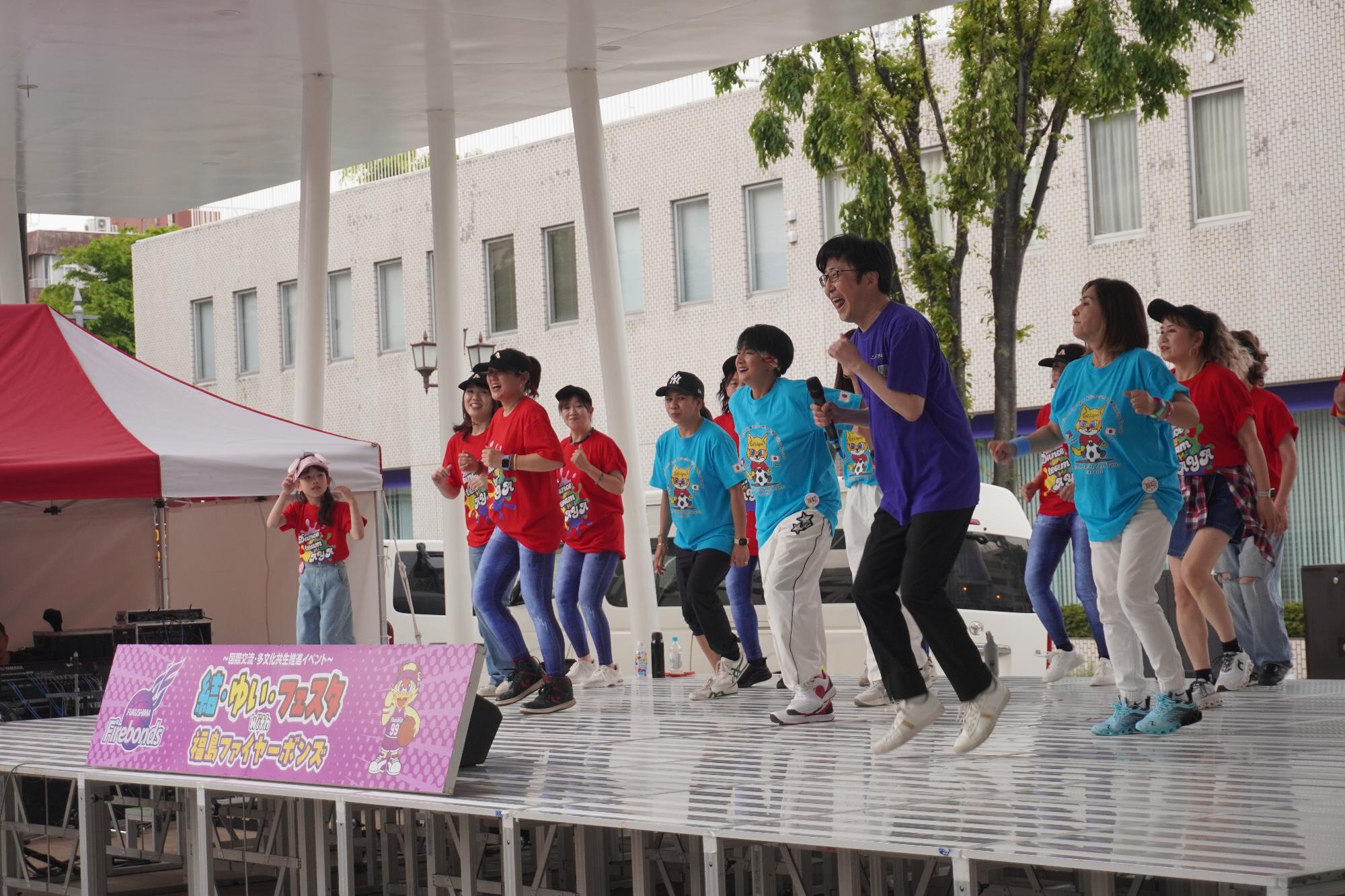 結・ゆい・フェスタイベントでのステージで、司会の引地さんと赤と青の服を着た団体みなさん合わせて10名程でエクササイズをしている様子