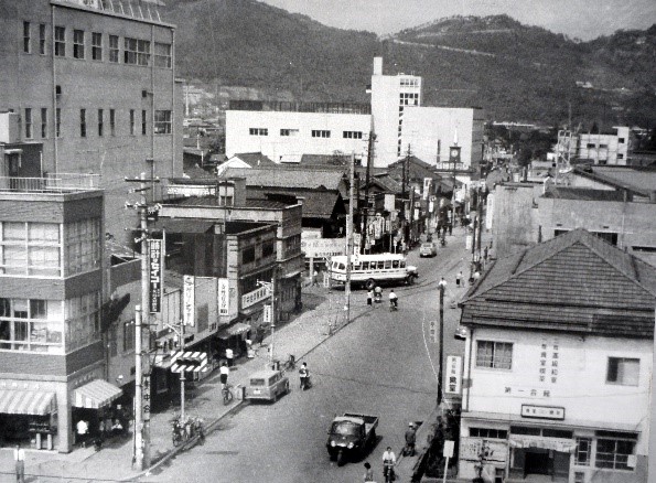 中央に幅の広い道路、両側にビルなどの建物が立ち、奥には山が見えている県庁通り商店街を写した白黒写真