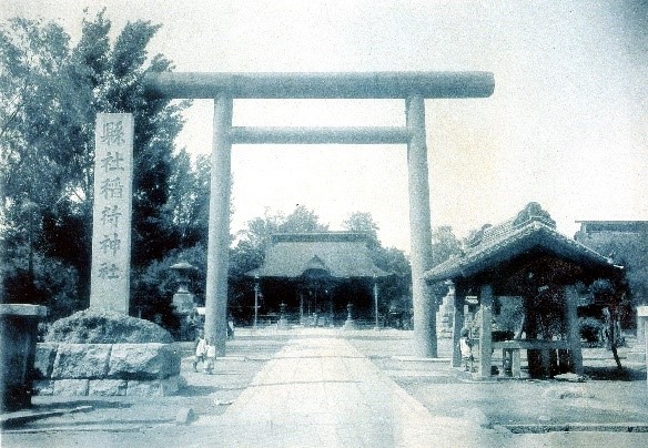 左側に「懸社稲荷神社」と書かれた大きな石柱、中央に鳥居、その奥に社殿が見えている稲荷神社を正面から写した白黒写真