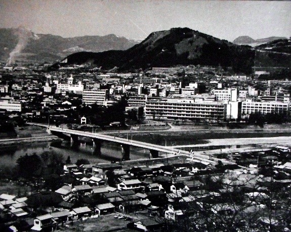 山の麓の大きな建物が立ち並んだ平地と、たくさんの住宅がたっている対岸を隔てる川にかかる天神橋を空撮した白黒写真