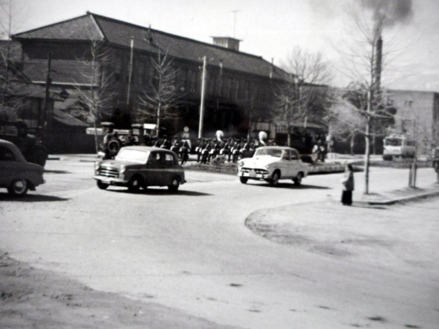 手前にカーブがかかった道路を3台の車が走っており、その奥に大きな2階建ての旧県立医科大学校舎が写っている白黒写真