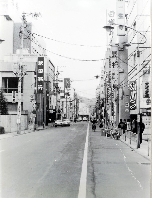 両側にビルなどが立ち並んでいる中央に1本の真っ直ぐな車道が通っている白黒写真