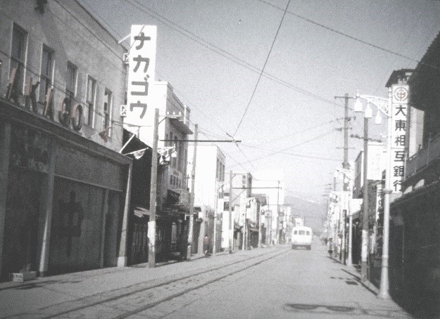 路面電車の路線がある一直線の道路の両側に建物が立ち並び、左手前の建物の2階部分には「ナカゴウ」の看板が設置されている様子を写した白黒写真