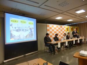 プロジェクタースクリーンが設置された横の長机の席に4名の方が座り、その横に男性1名が起立し出版発表記者会見が行われている様子の写真