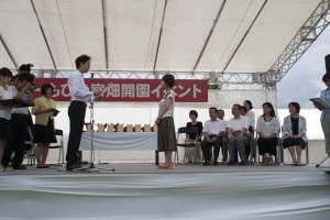 スタンドマイクの前に男性が立ち、その向かい側に女性が立ち表彰式が行われている様子の写真