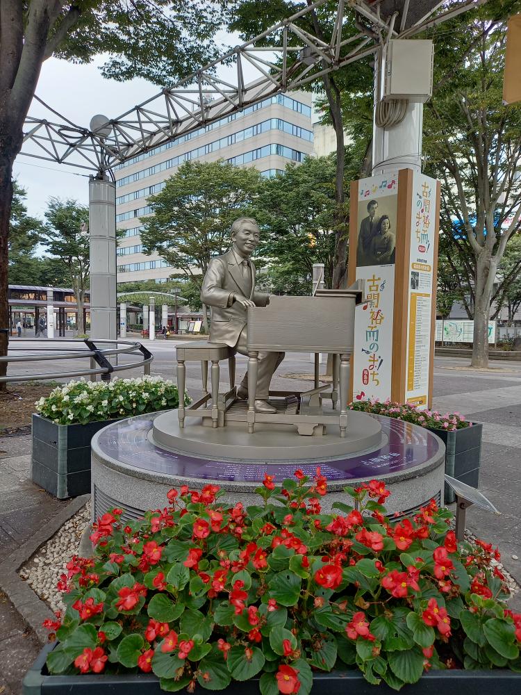 花壇に囲まれた円柱状の土台の上にあるピアノを弾いている様子の古関裕而氏のモニュメントの写真