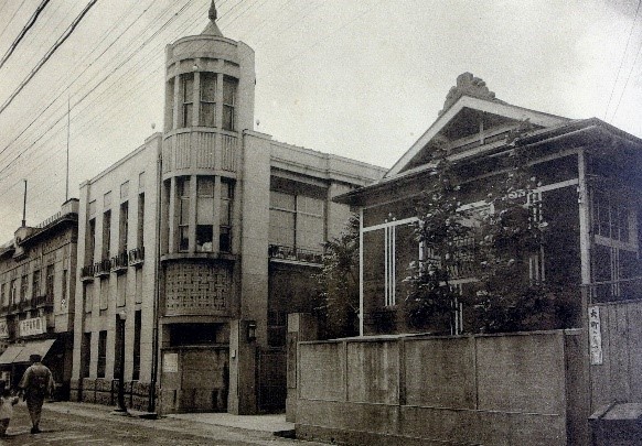 左側に2階建てで角が丸みを帯びたコンクリート製のような建物、右側に三角屋根で平屋建ての建物が写っている白黒写真